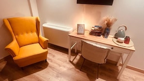 A room with a wooden floor, a yellow armchair, a desk with a lamp, books, a teapot, and a vase.