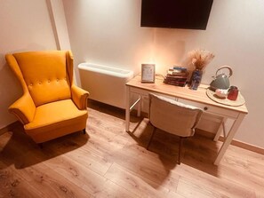 Una habitación con suelo de madera, un sillón amarillo, un escritorio con lámpara, libros, una tetera y un jarrón.