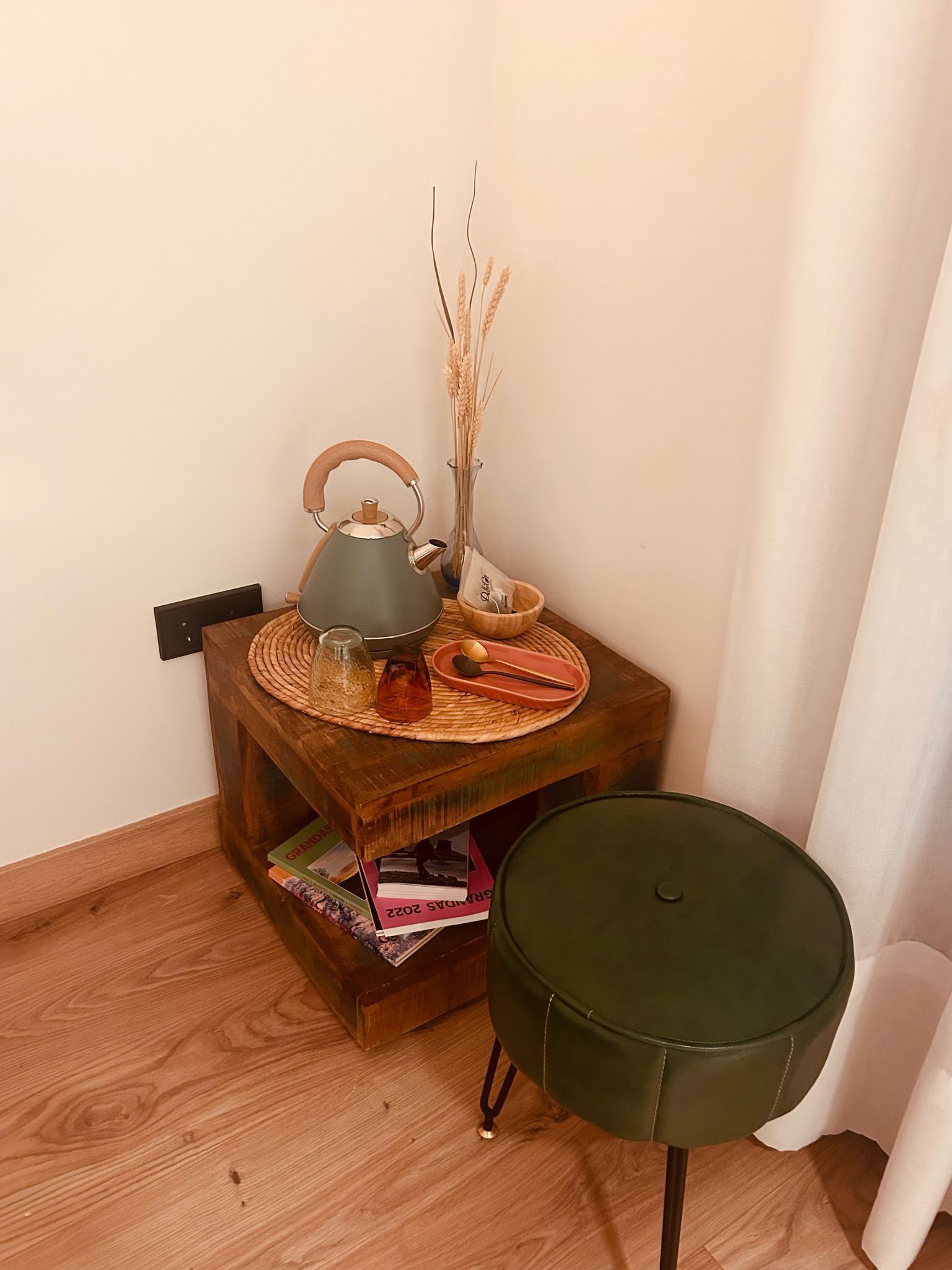 A corner of a room with a wooden table, a green chair, a kettle, a vase with dried plants, and a stack of books.