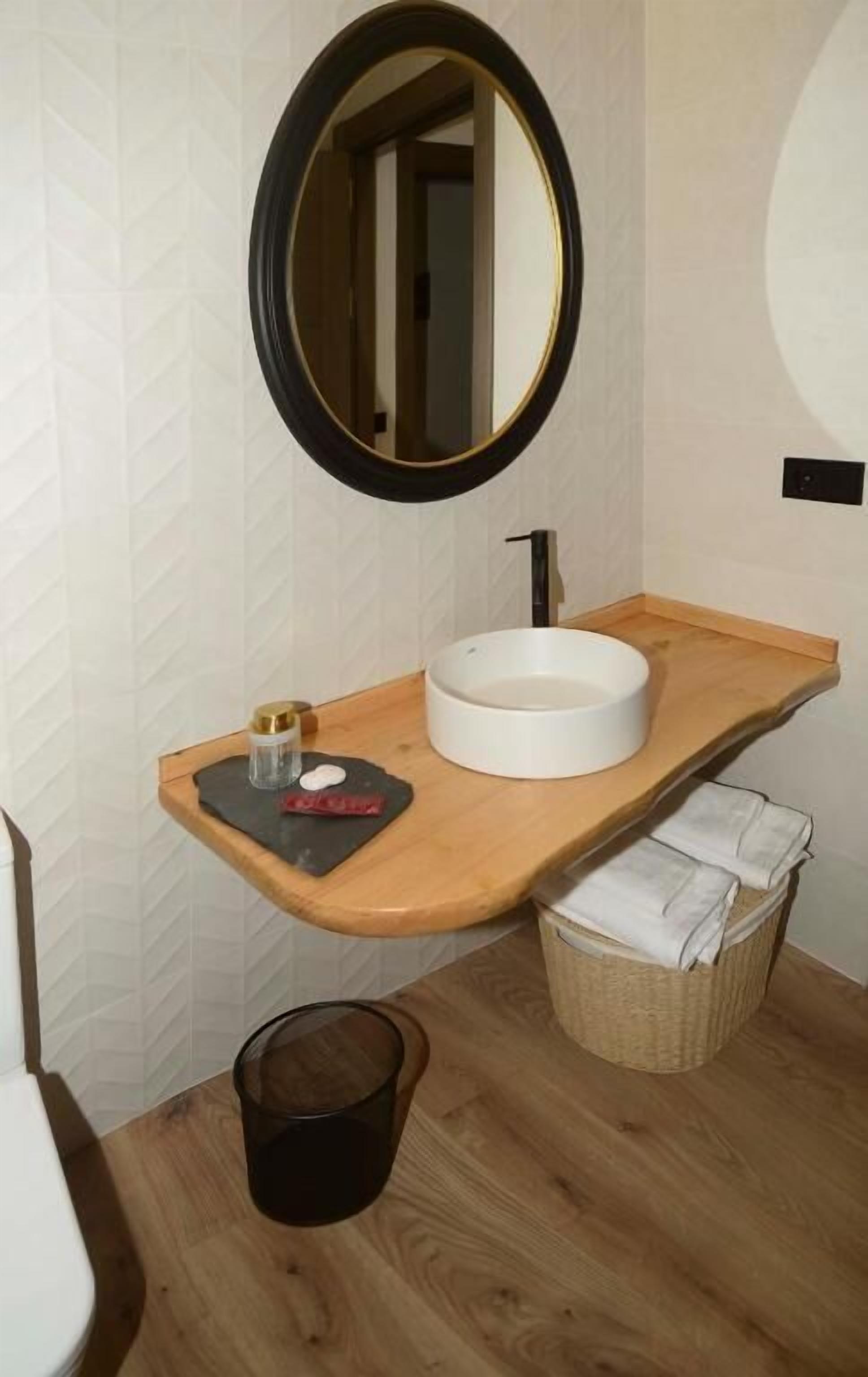 Une salle de bain avec un meuble-lavabo en bois, un lavabo rond, un miroir et une petite poubelle.