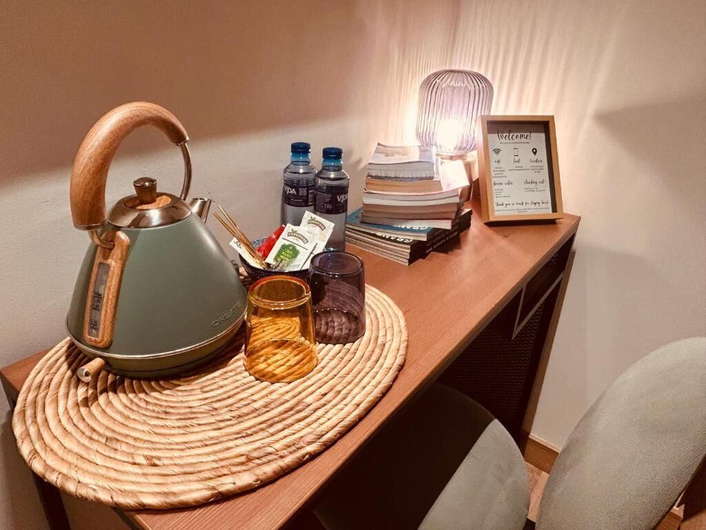 Un escritorio de madera con un camino de mesa tejido, una tetera verde, dos botellas de agua, una pila de libros, un cartel de bienvenida en un marco y un candelabro.