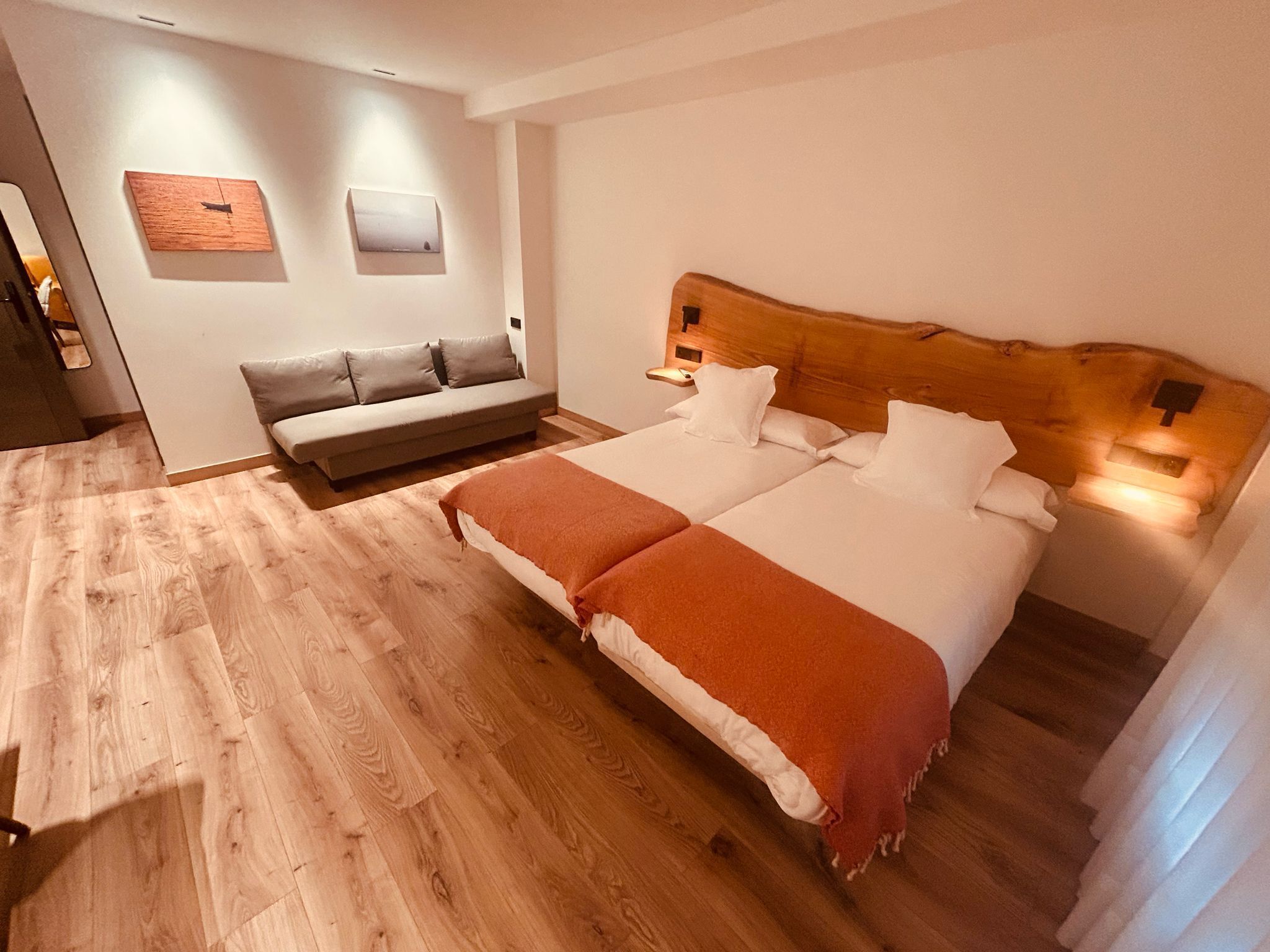 A bedroom with a wooden headboard, a double bed with white bedding, a wooden floor, a grey sofa, and a wall-mounted light fixture.