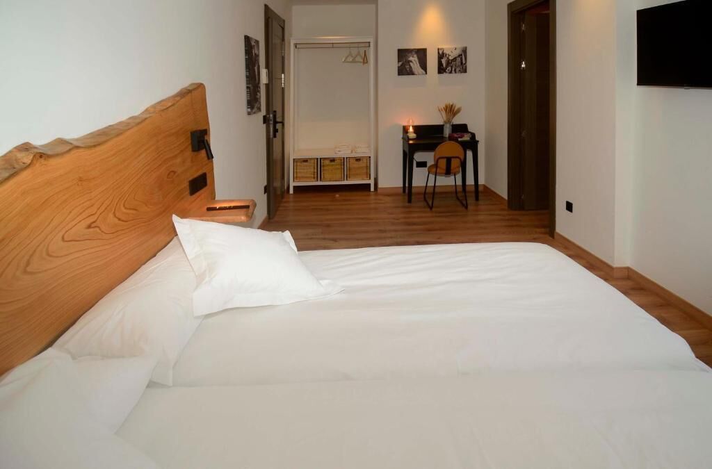 Un dormitorio con cabecera de madera, cama con ropa de cama blanca, escritorio con silla y televisor montado en la pared.