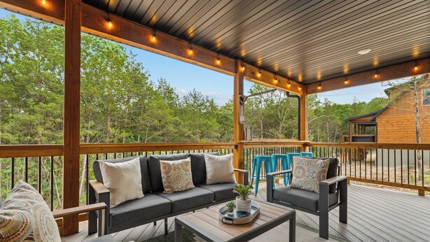 An outdoor deck with a wooden bench, a sofa, and a coffee table, surrounded by a railing and trees. - HotTub+Arcade+Pool | 15min to Dolly's Stampede (Hollister)