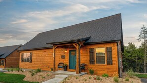 A two-story log cabin with a dark shingle roof, a front porch with two chairs, and a green door. - HotTub+Arcade+Pool | 15min to Dolly's Stampede (Hollister)
