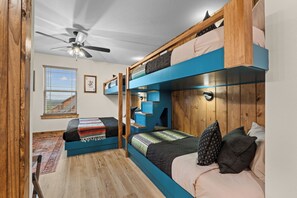 Ein kleines Zimmer mit Etagenbetten, einem Deckenventilator, einem Fenster mit Jalousien und einer gerahmten Spielkarte an der Wand.