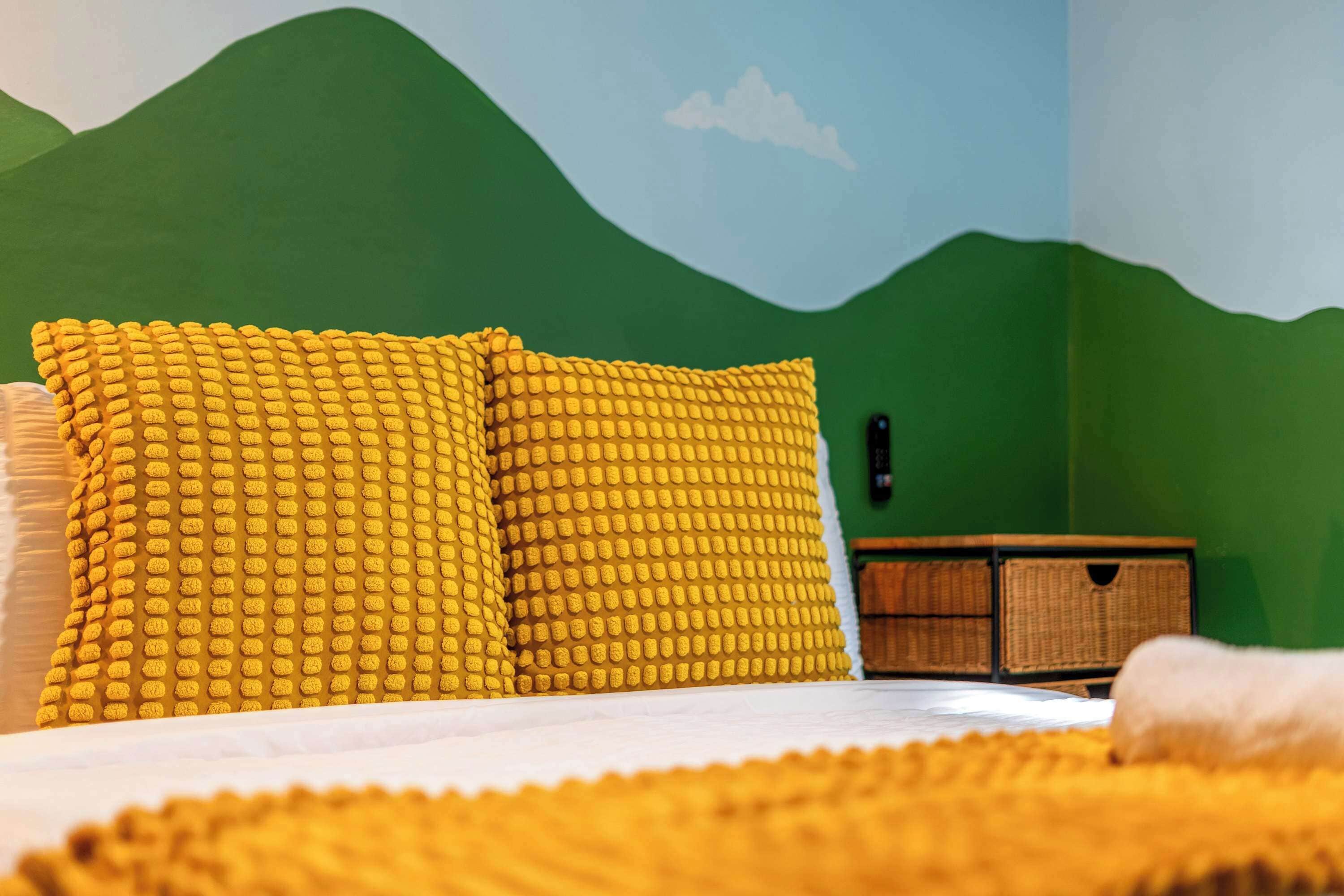Ein Bett mit gelben, strukturierten Kissen und einer weißen Decke, vor einer grünen Wand mit Bergmotiv.