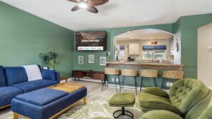 A living room with a sofa, ottoman, and green chairs, a kitchen area with a bar, and a TV displaying 'Welcome to Aloha Springs'. - Aloha Springs! Private Heated Pool & Hot Tub (Colorado Springs)