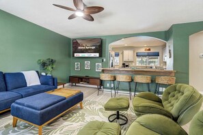 A living room with a sofa, ottoman, and green chairs, a kitchen area with a bar, and a TV displaying 'Welcome to Aloha Springs'. - Aloha Springs! Private Heated Pool & Hot Tub (Colorado Springs)