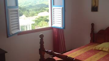A bedroom with a bed, a window with shutters, a painting on the wall, and a view of greenery outside.