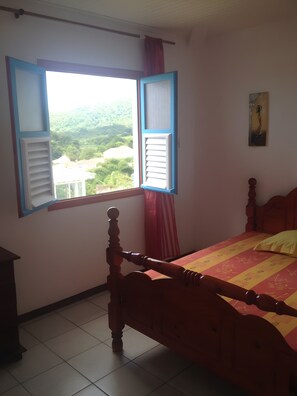 A bedroom with a bed, a window with shutters, a painting on the wall, and a view of greenery outside. - Guest House 'Villa Pelée' with Sea View, Balcony and Wi-Fi (Le Morne-Rouge)