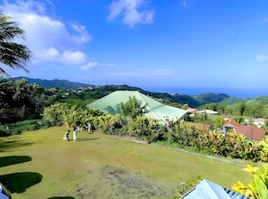 A grassy area with a green tent-like structure, surrounded by lush vegetation and houses in the background. - Guest House 'Villa Pelée' with Sea View, Balcony and Wi-Fi (Le Morne-Rouge)