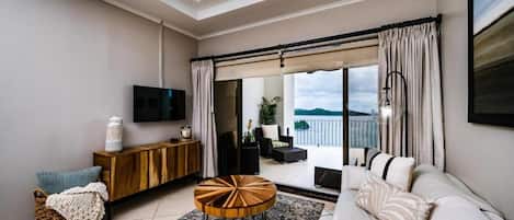 Una sala de estar moderna con un sofá, una mesa de centro redonda y un televisor. Hay un balcón con vistas al cielo y al agua.
