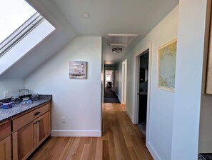 A narrow hallway with wooden flooring, a kitchen area on the left with granite countertops, and a dining area visible through an open door. - Luxury Coastal Apt Above Bakery,Near Beach, Acadia (Hancock)
