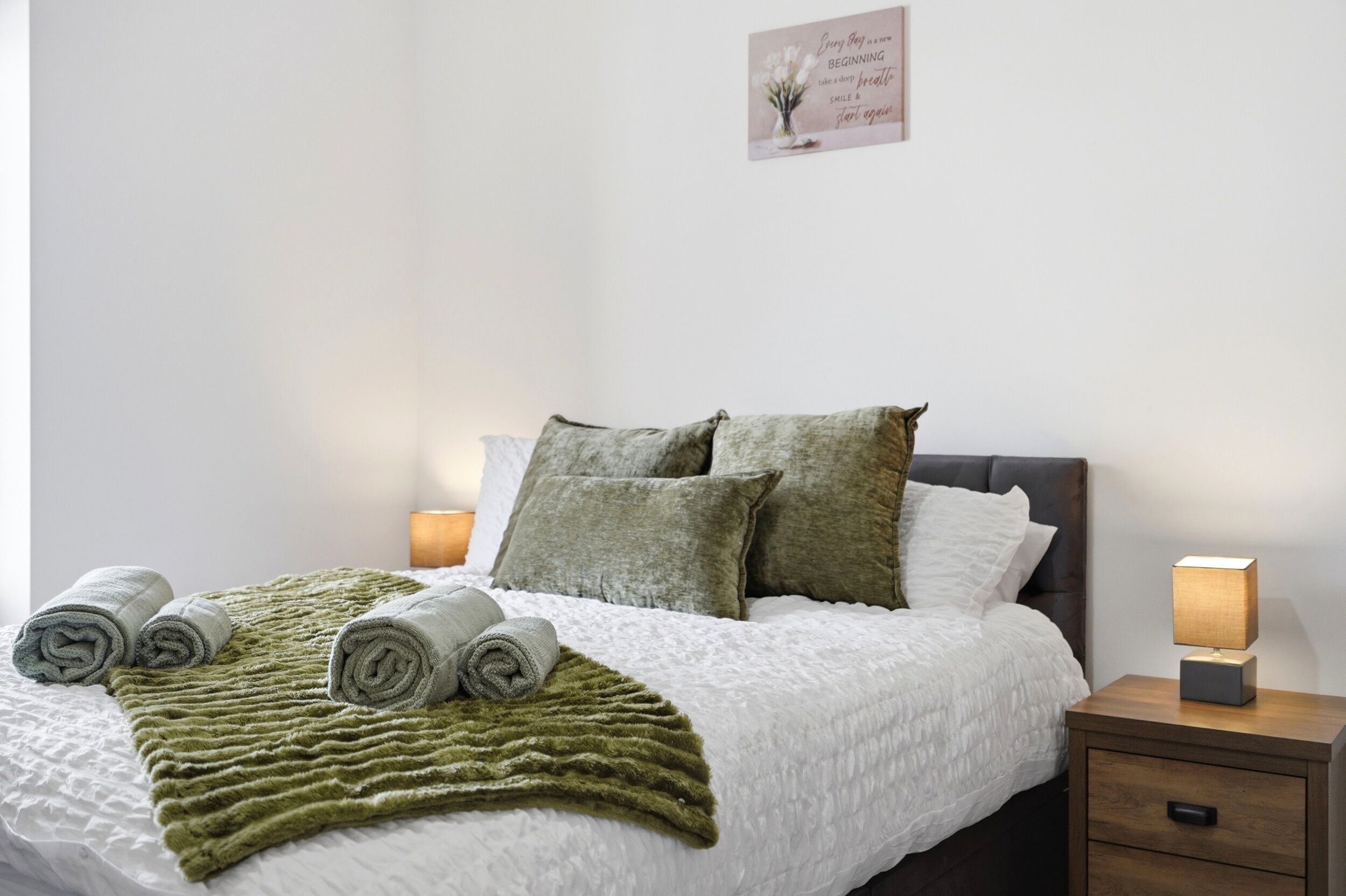 A neatly made bed with rolled towels, a green blanket, and two pillows on a white duvet. There is a bedside table with a lamp and a small drawer. A motivational quote is displayed on the wall above the bed.