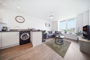 A modern apartment with a kitchen, a living area with a sofa and a coffee table, a washing machine, and a large window with blinds. - Comfortable Stay | Fast Wi-Fi | Near Train Station | Sleeps 2 (Coventry)