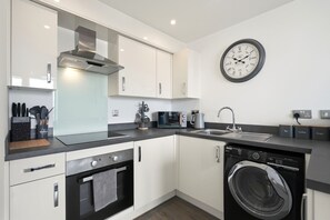 A modern kitchen with white cabinets, a built-in oven, a microwave, a coffee machine, and a washing machine. - Comfortable Stay | Fast Wi-Fi | Near Train Station | Sleeps 2 (Coventry)