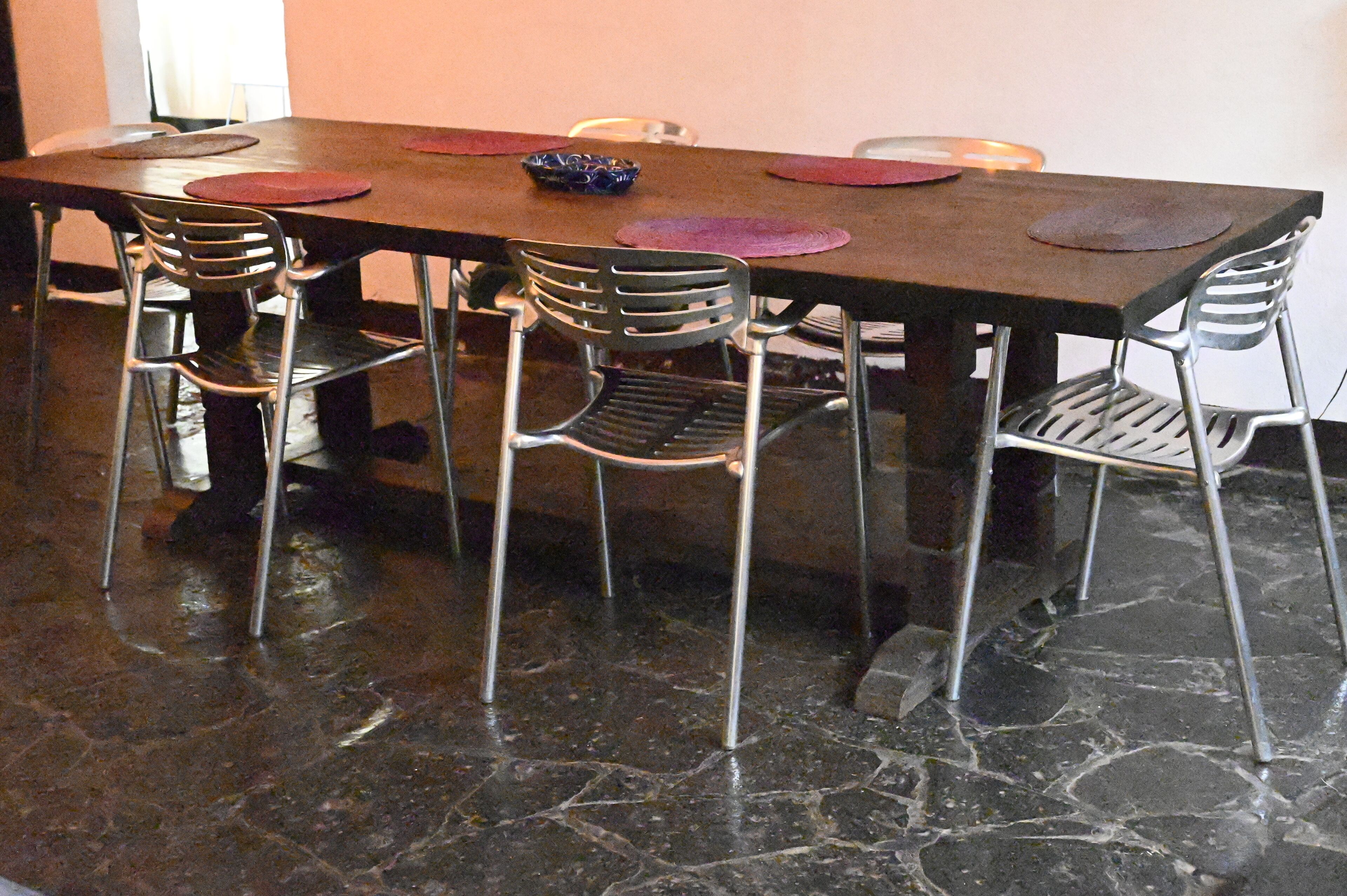 Una mesa rectangular de comedor con seis sillas, cada una con estructura metálica y respaldo de malla. La mesa está cubierta con un mantel oscuro, y hay dos platos decorativos sobre ella.
