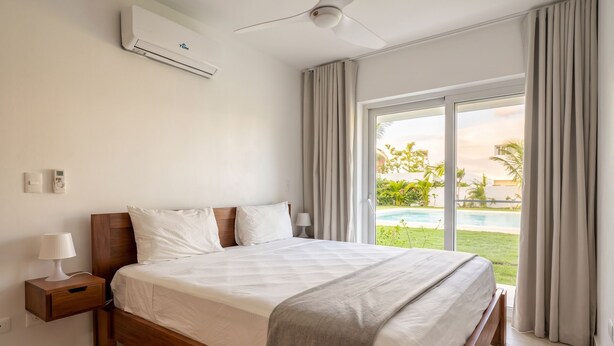 Ein Hotelzimmer mit Bett, Nachttisch mit Lampe, Klimaanlage und Blick auf einen Pool.
