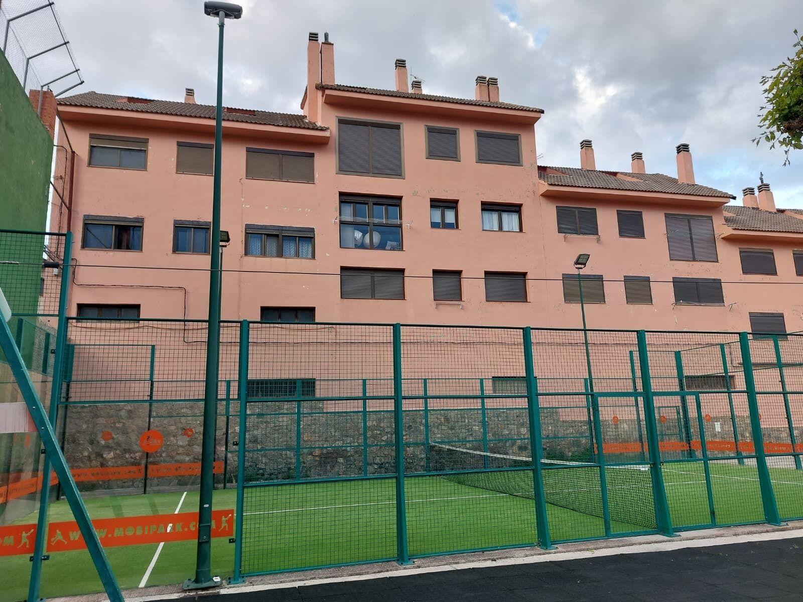 A multi-story building with a sports field enclosed by a green fence.