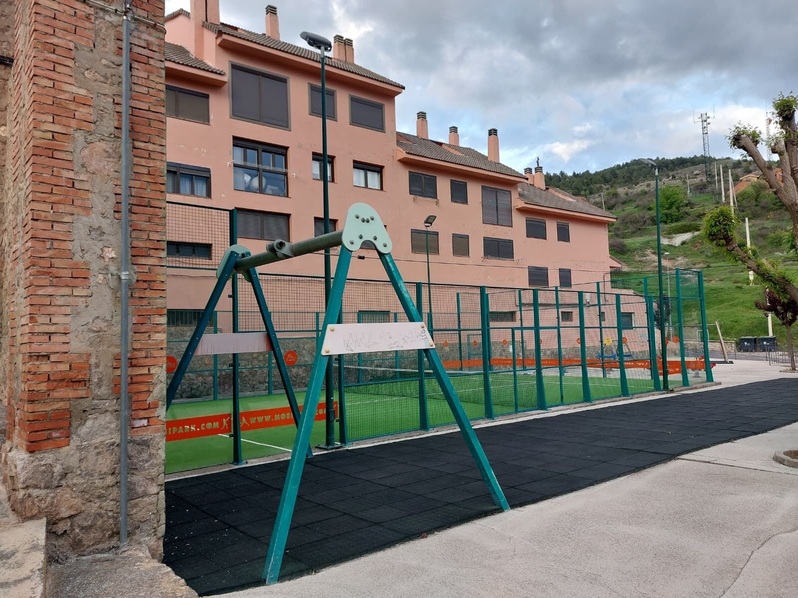 Un parque infantil con columpios y un área cercada, un muro de ladrillo y un edificio con varias ventanas al fondo.