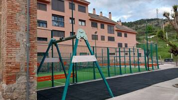 Un parque infantil con columpios y un área cercada, un muro de ladrillo y un edificio con varias ventanas al fondo.