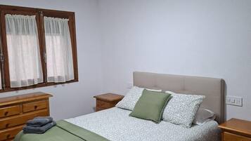 Un dormitorio con una cama, dos mesitas de noche, una ventana con cortinas e iluminación en el techo.