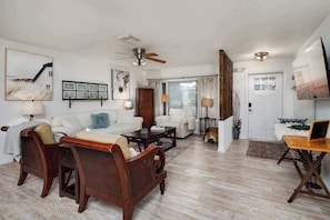 A spacious living room with a sofa, coffee table, and a wooden chair. There is a ceiling fan, a fireplace, and a door leading to another room.