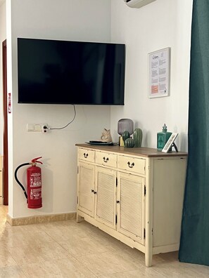 Een kamer met een wandgemonteerde flatscreen-tv, een brandblusser, een kast met lades en een cactus er bovenop.