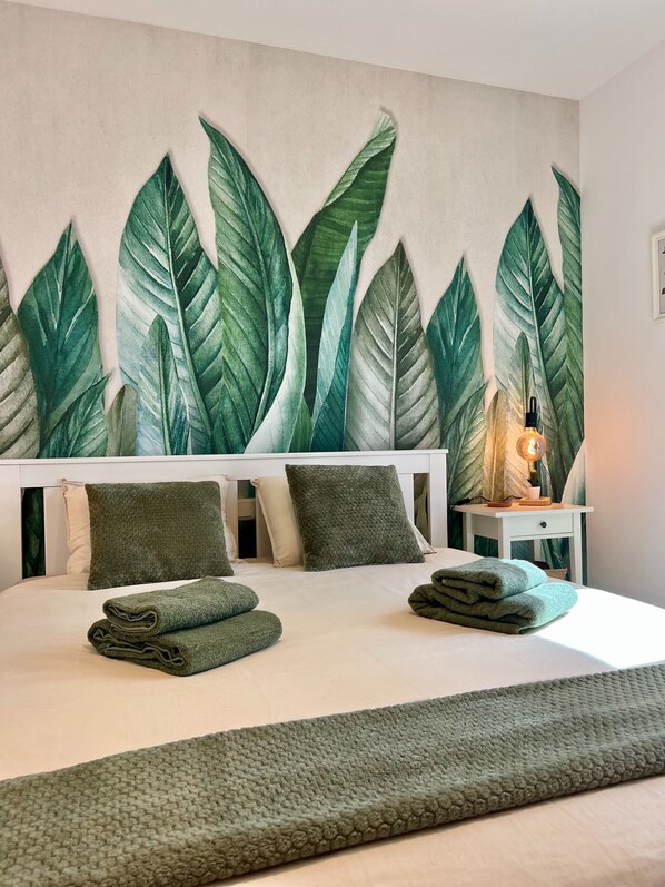 Quarto com uma cama grande, duas almofadas, uma mesa de cabeceira com abajur e uma parede com padrão de folhagem.