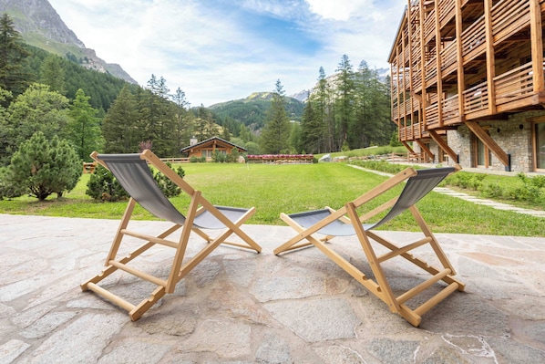 Two wooden lounge chairs on a stone patio with a view of a mountain and a wooden building in the background.