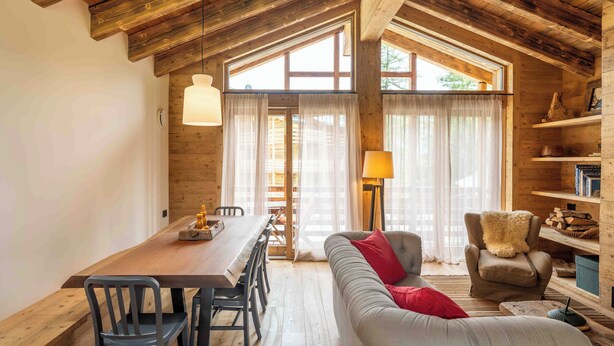 A cozy interior with a wooden ceiling, a dining area with a table and chairs, a sofa with a red pillow, and a bookshelf.