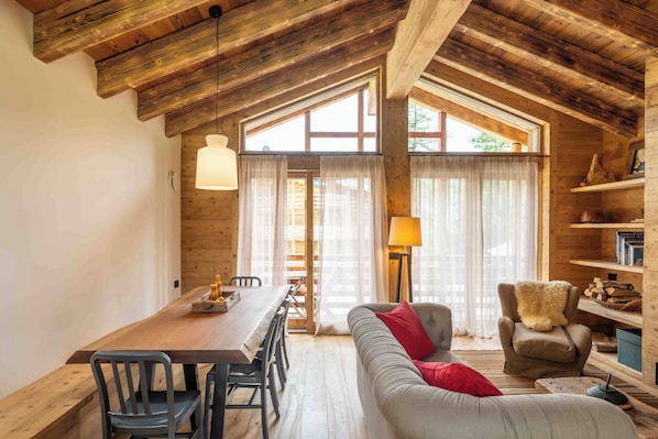 A cozy interior with a wooden ceiling, a dining area with a table and chairs, a sofa with a red pillow, and a bookshelf.