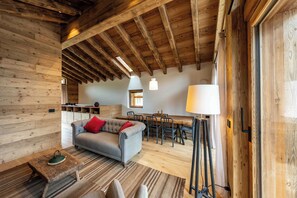 A rustic interior with exposed wooden beams, a grey sofa with red cushions, a dining area with a wooden table and chairs, and a large window providing natural light.