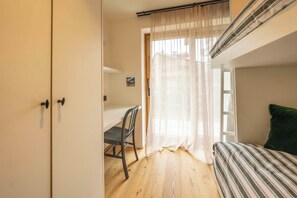 A bedroom with a bed, a chair, a desk, and a window with sheer curtains.