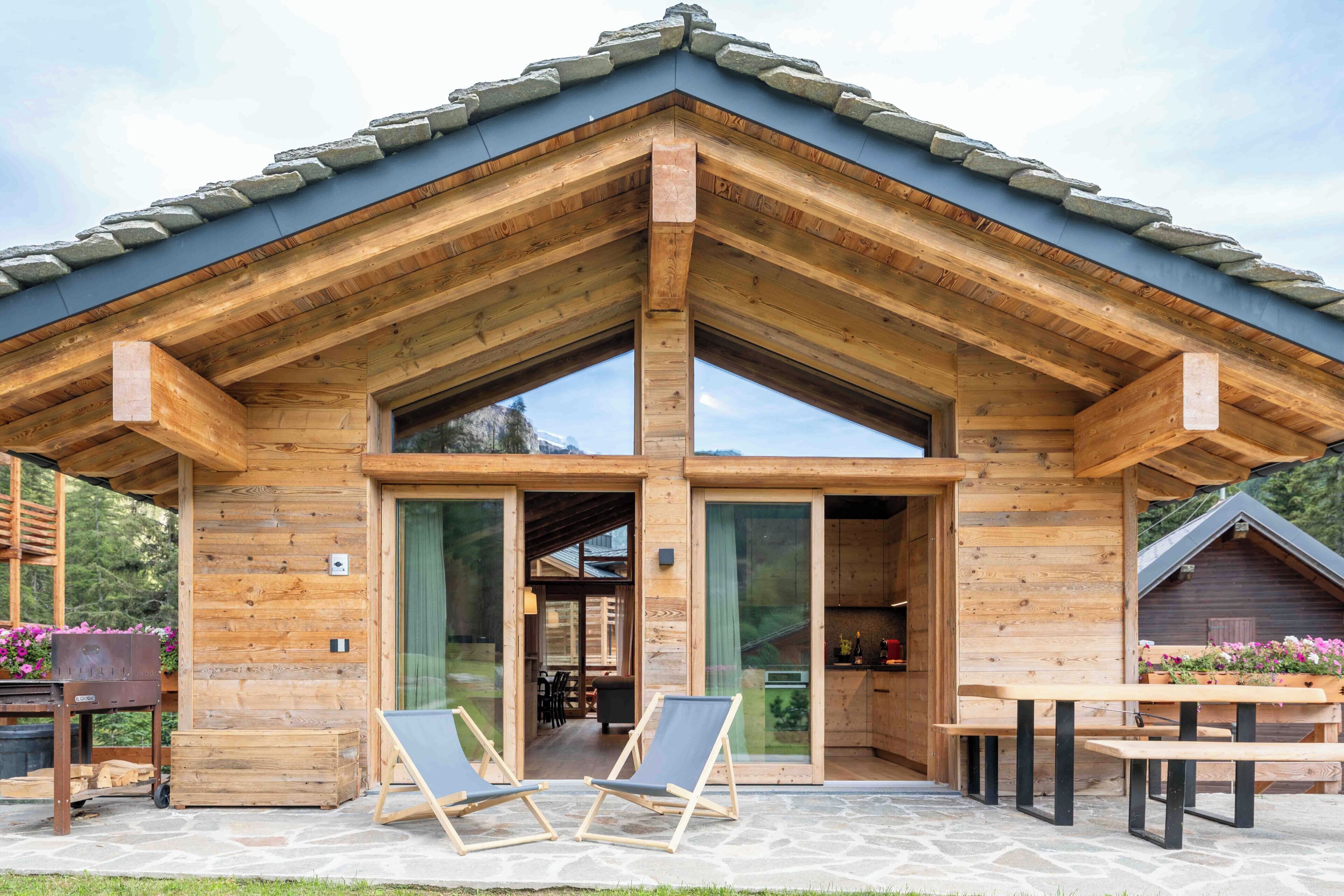 Une cabane en bois avec un toit en pente, de grandes portes vitrées et un patio en pierre doté de meubles d’extérieur.