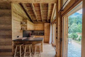 A wooden interior with a kitchen area, stools, and a view of the outdoors.