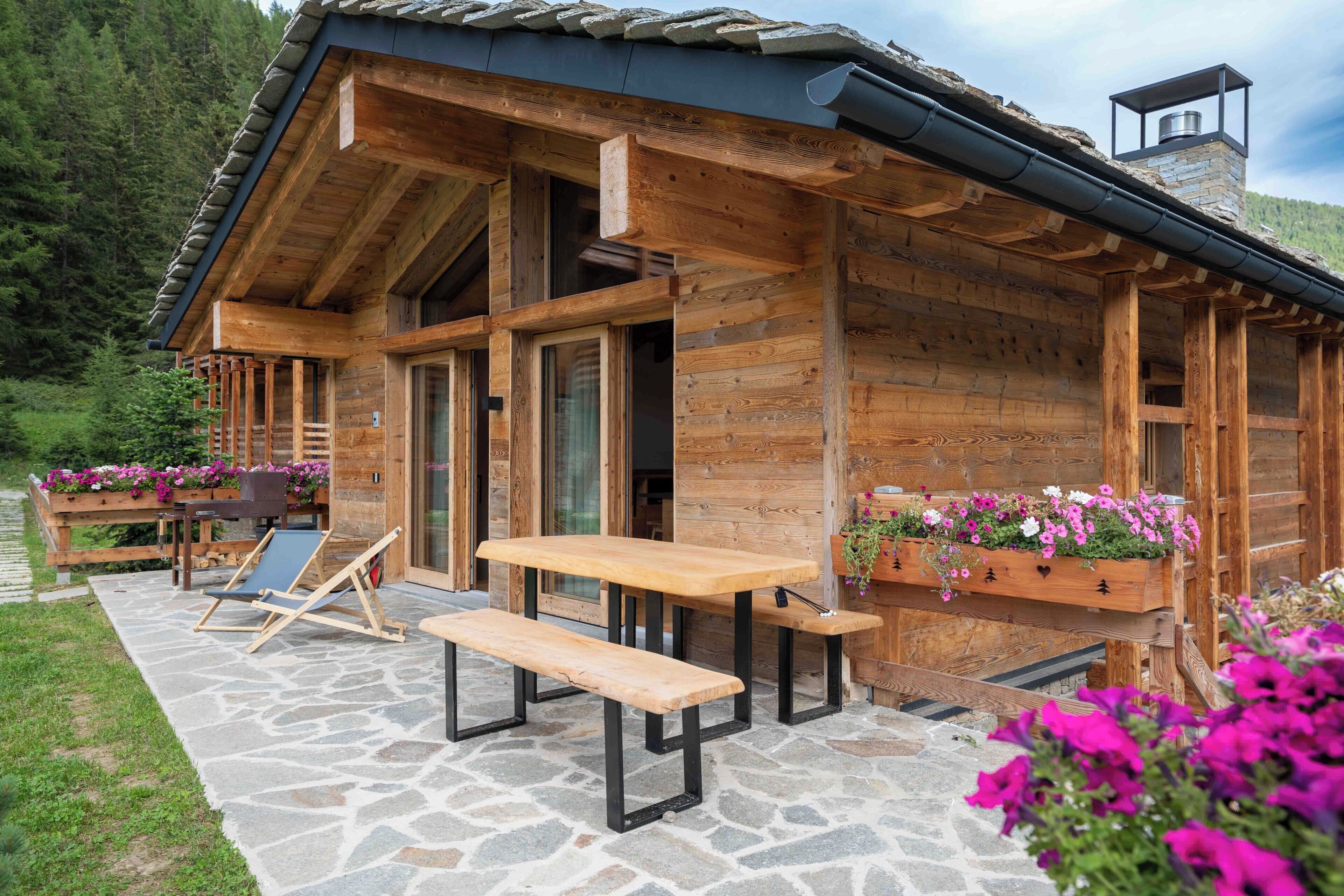 A wooden cabin with a stone patio, outdoor seating, and flower planters.