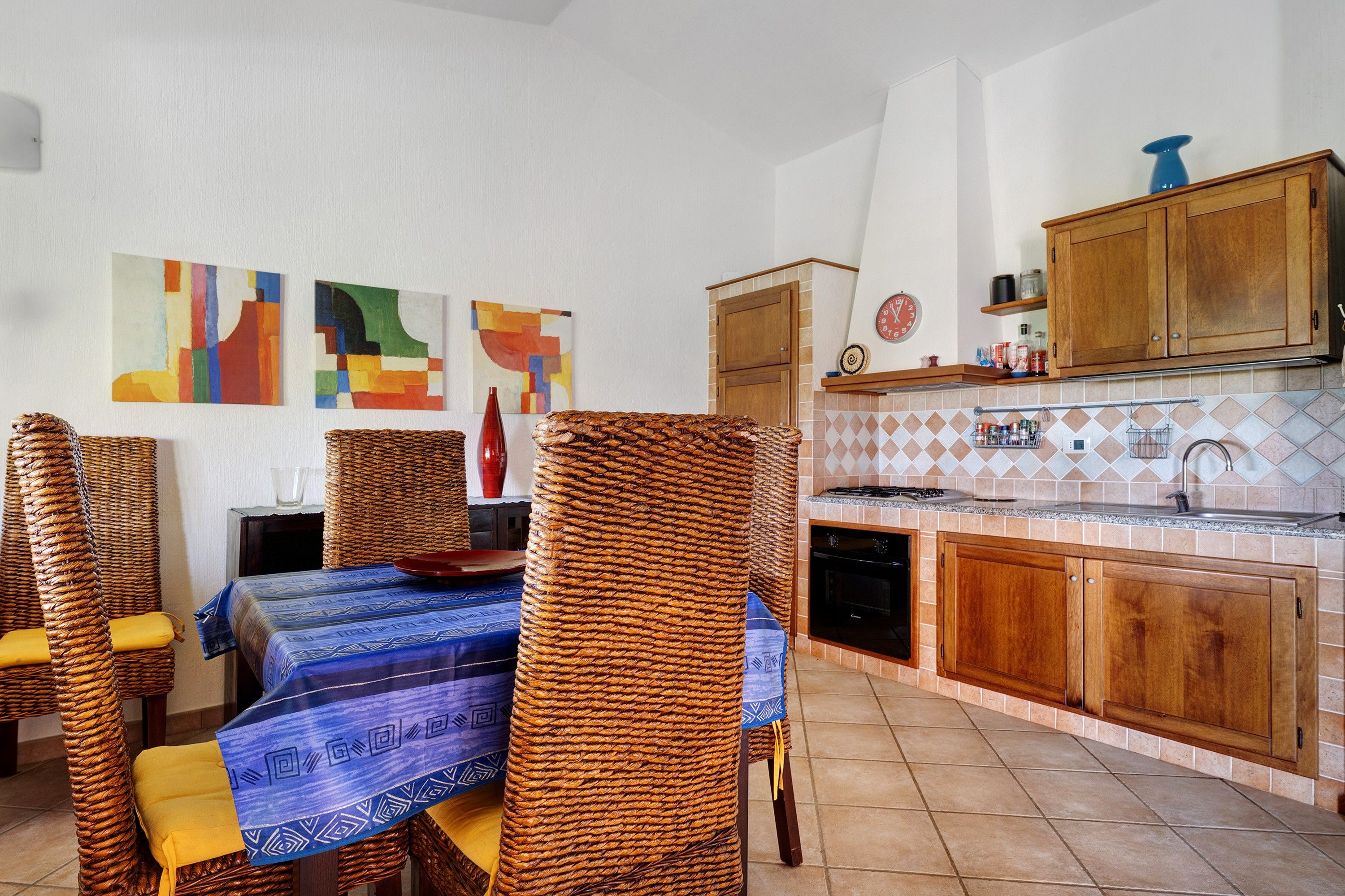 Un coin repas avec des chaises en osier, une table recouverte d’une nappe à motifs bleus, et une cuisine dotée de meubles en bois et d’un dosseret carrelé.