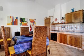 Ein Essbereich mit Korbstühlen, einem Tisch mit blau gemustertem Tischläufer und einer Küche mit Holzschränken und einer gefliesten Spritzschutzleiste.