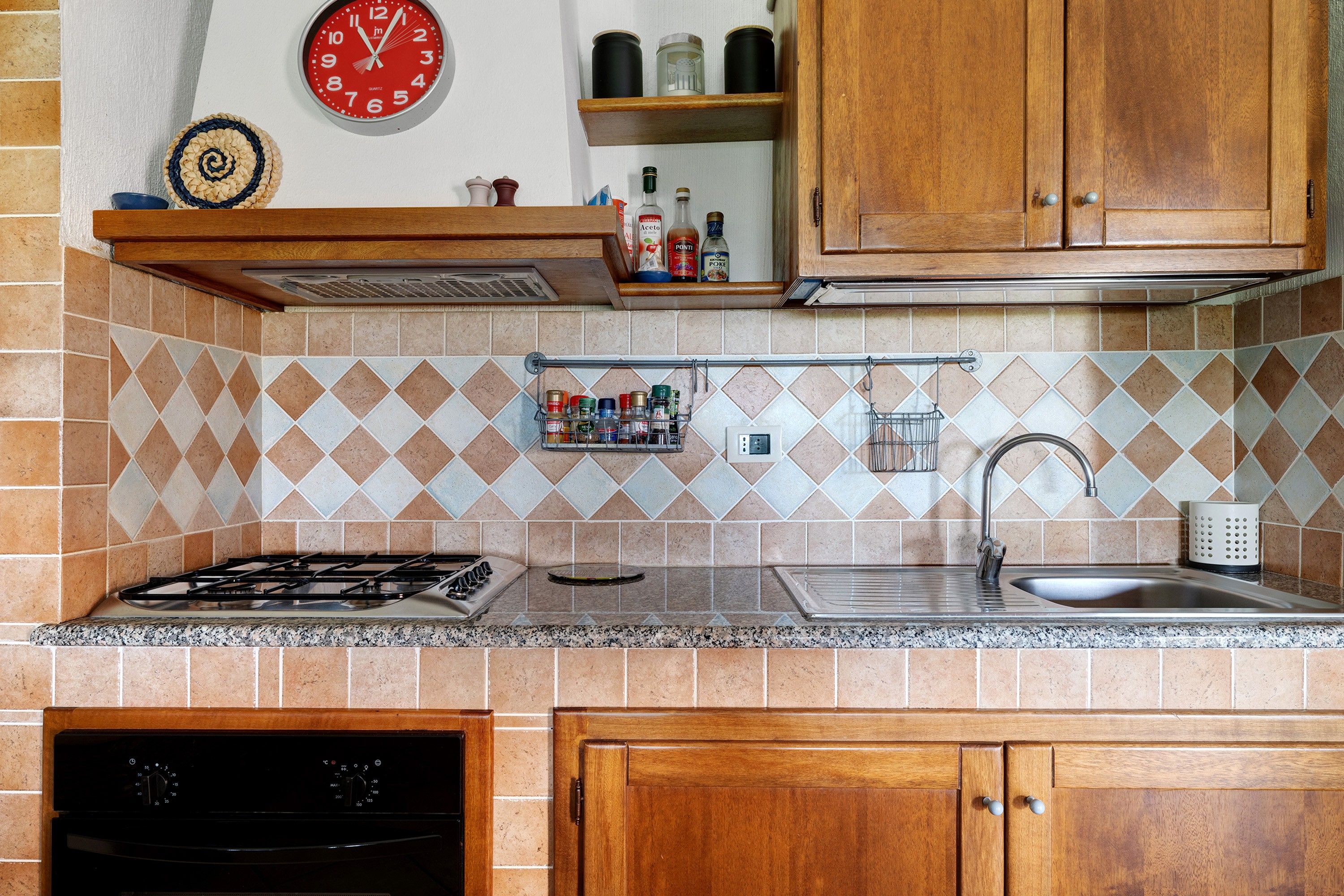 A kitchen with a gas stove, a sink, wooden cabinets, a spice rack, and a wall clock.