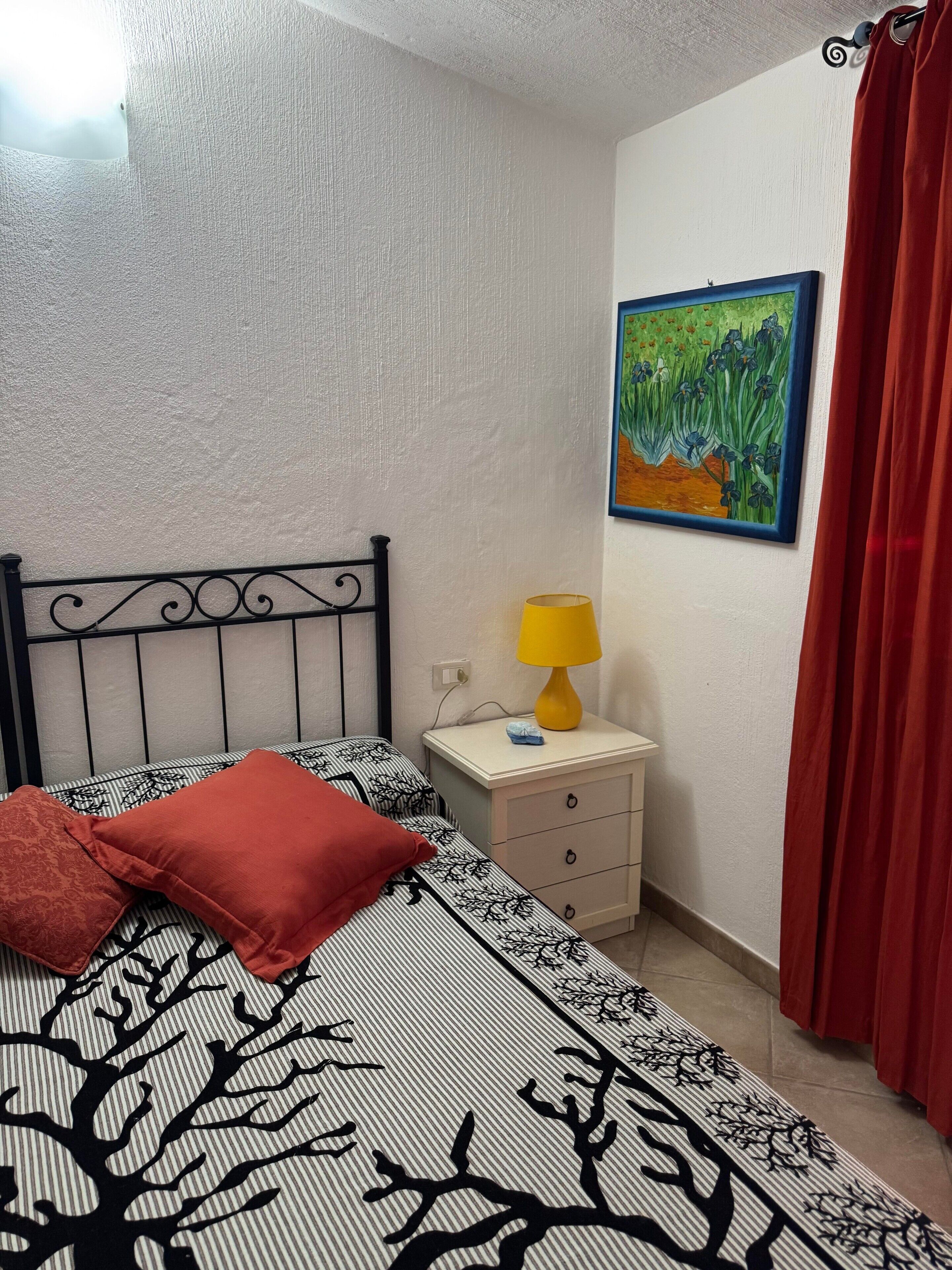 A bedroom with a bed, a nightstand with a lamp, a painting on the wall, and a red curtain.