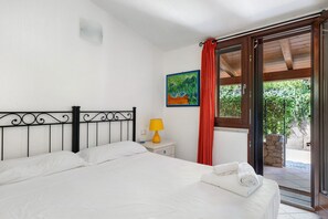 A bedroom with a bed, a nightstand, a lamp, a painting, and a view of the outdoors. - Villino Empatja: an emerald sea at your doorstep (Tertenia)