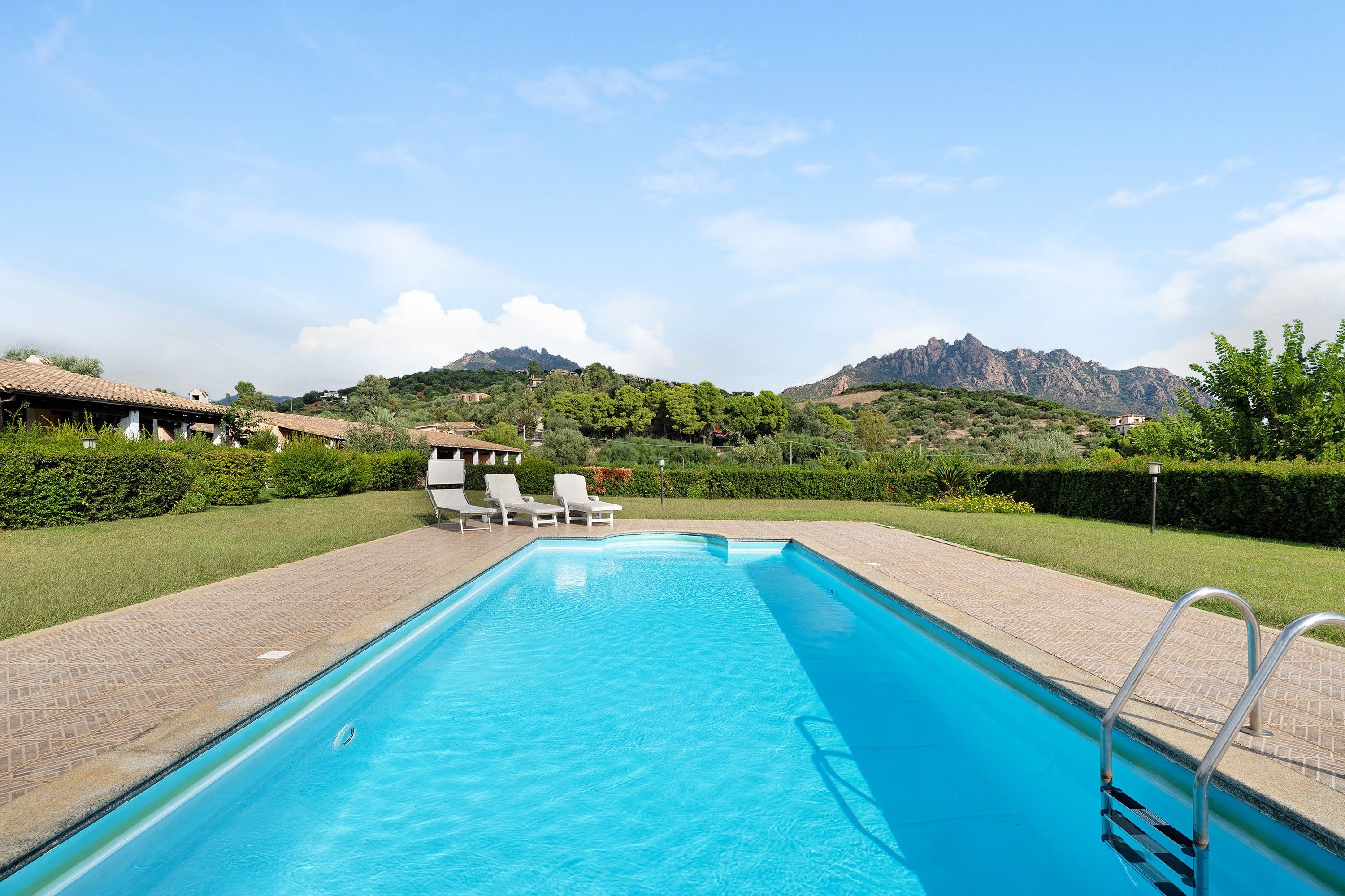 Ein rechteckiger Swimmingpool mit klarem, blauem Wasser, umgeben von einer gepflasterten Liegefläche. Auf der Liegefläche stehen Liegestühle und eine Poolleiter. Im Hintergrund sind Hügel und ein Gebirge zu sehen.