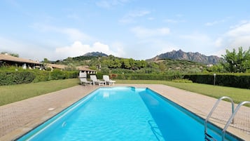 A rectangular swimming pool with clear blue water, surrounded by a paved deck. There are lounge chairs and a pool ladder on the deck. In the background, there are hills and a mountain range.