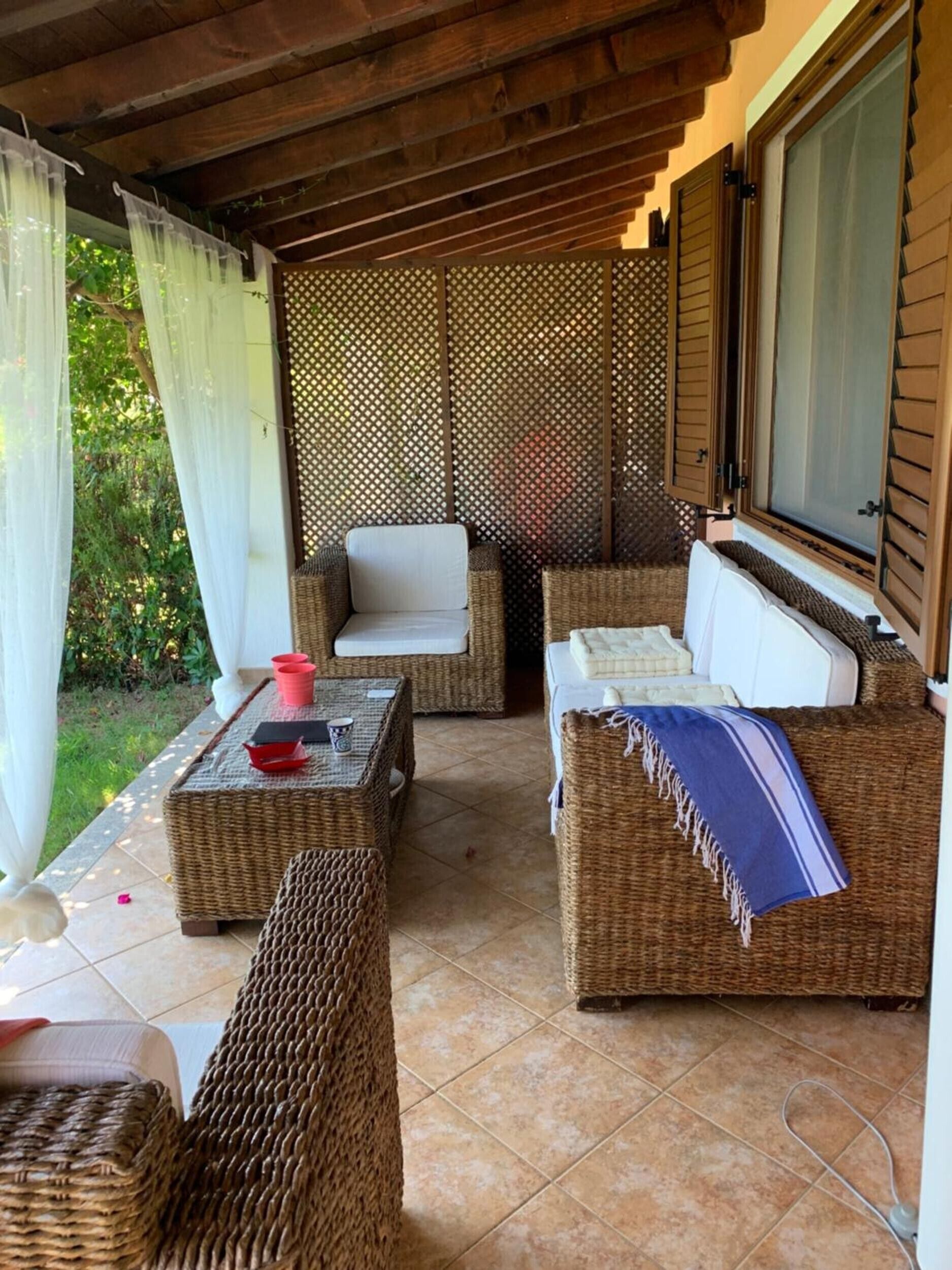 A covered outdoor seating area with wicker furniture, a table with cups, and a view of greenery.