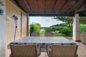 Eine Terrasse im Freien mit Tisch und Stühlen, ein überdachter Bereich mit Holzboden und Blick auf einen Garten und Hügel im Hintergrund.