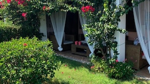 A covered outdoor seating area with white curtains and a garden with green shrubs and red flowers.