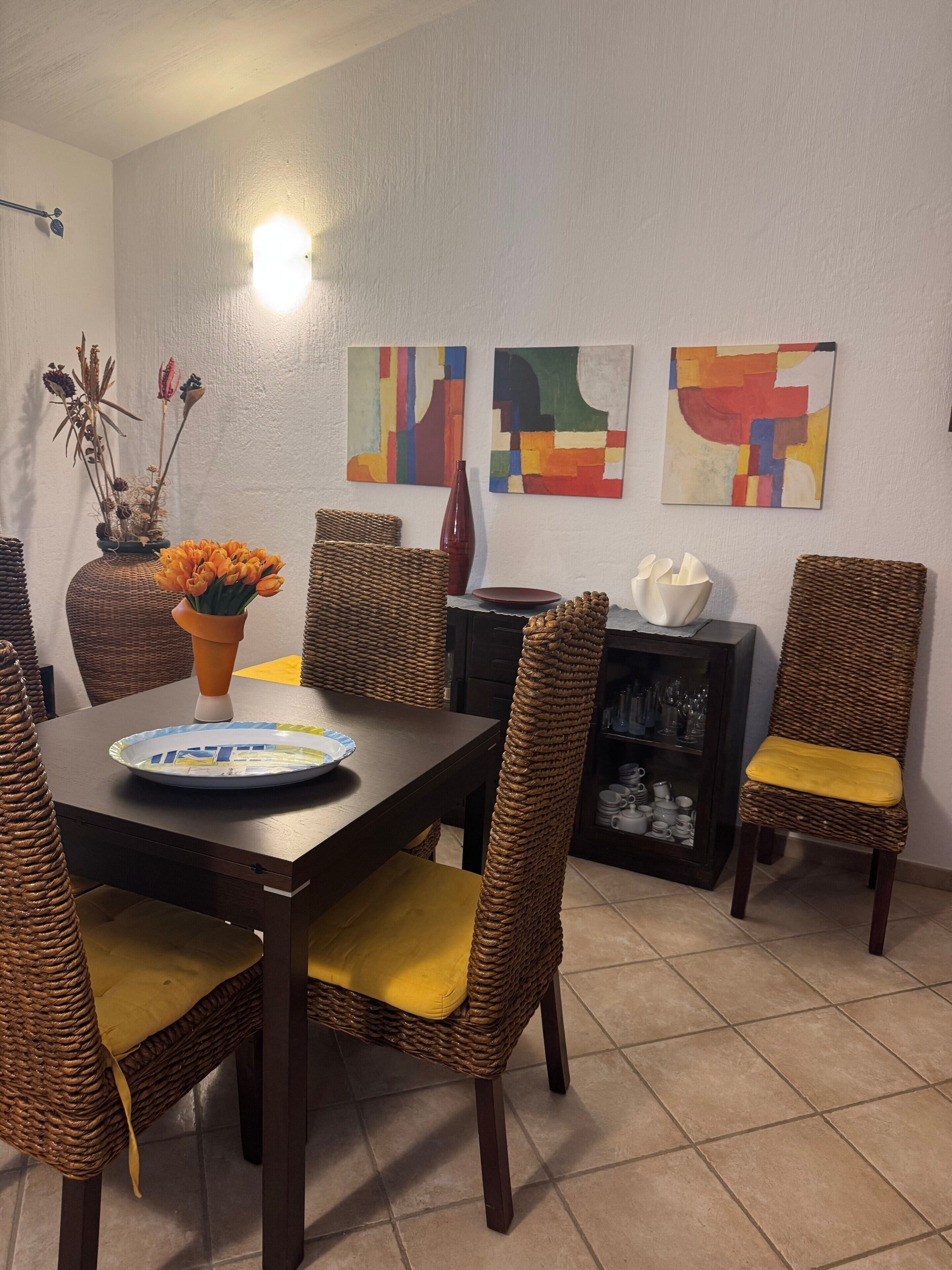 A dining area with a dark wooden table, wicker chairs with yellow cushions, and a vase with orange flowers. There are abstract paintings on the wall and a shelf with glassware.
