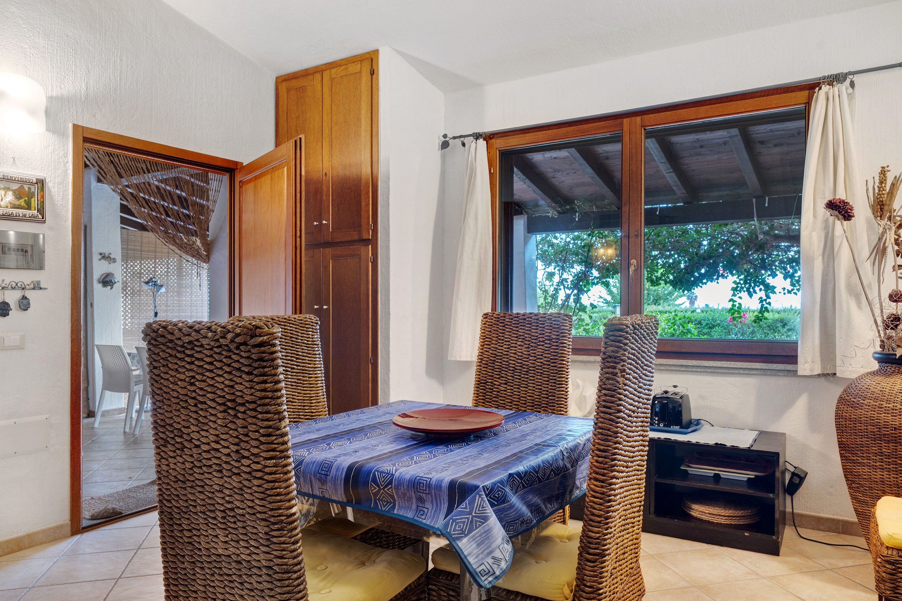 A dining area with a table set for two, surrounded by wicker chairs, a wooden door, and a window with a view of greenery.
